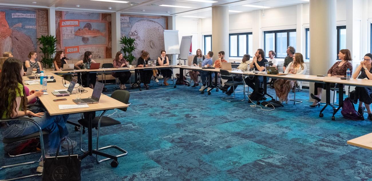 A room with tables in a u-shaped setting. People are sitting at the tables listing/looking to one person that speaks